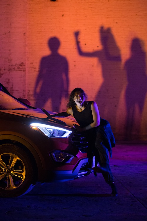 A woman dances on the hood of a car, her mouth agape. 